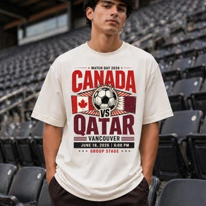 May include: Cream-colored t-shirt with a soccer-themed graphic. The design features the word "CANADA" in red above a soccer ball and the word "QATAR" in maroon. The text "VANCOUVER JUNE 18, 2026 6:00 PM GROUP STAGE" is also included.