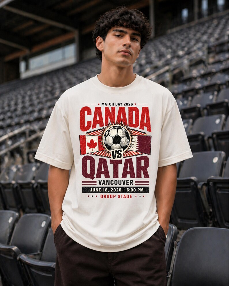 May include: Cream-colored t-shirt with a soccer-themed graphic. The design features the word "CANADA" in red above a soccer ball and the word "QATAR" in maroon. The text "VANCOUVER JUNE 18, 2026 6:00 PM GROUP STAGE" is also included.