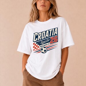 以下が含まれることがあります： CROATIA Soccer Club 26 NORTH AMERICA 26と書かれたグラフィックプリントが施された白い半袖Tシャツ。デザインには、赤と白の市松模様の正方形、サッカーボールのイラスト、赤と青の水平線が含まれています。