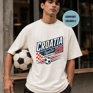 以下が含まれることがあります： 「CROATIA Soccer Club 26」および「NORTH AMERICA 26」の文字がプリントされたオフホワイトの半袖Tシャツ。赤と白の市松模様、サッカーボールのイラスト、赤と青の水平線がデザインされています。