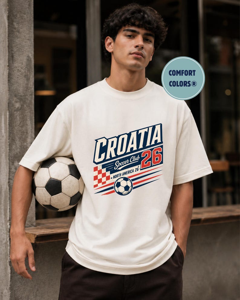 以下が含まれることがあります： 「CROATIA Soccer Club 26」および「NORTH AMERICA 26」の文字がプリントされたオフホワイトの半袖Tシャツ。赤と白の市松模様、サッカーボールのイラスト、赤と青の水平線がデザインされています。