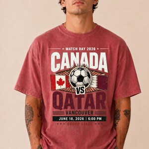 May include: Red t-shirt with a graphic design for the Canada vs. Qatar soccer match in Vancouver, June 18, 2026. The design includes the flags of Canada and Qatar, a soccer ball, and the text "CANADA" and "QATAR".