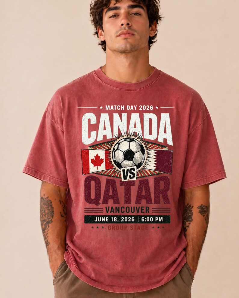May include: Red t-shirt with a graphic design for the Canada vs. Qatar soccer match in Vancouver, June 18, 2026. The design includes the flags of Canada and Qatar, a soccer ball, and the text "CANADA" and "QATAR".