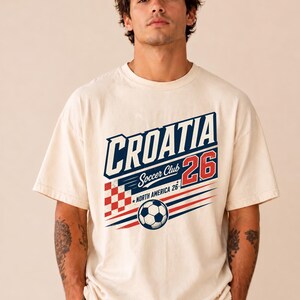 以下が含まれることがあります： CROATIA、Soccer Club、NORTH AMERICA 26という文字がプリントされたクリーム色の半袖Tシャツ。デザインには、赤と白のチェック柄の四角形、赤色の数字26、そして白黒のサッカーボールが含まれています。