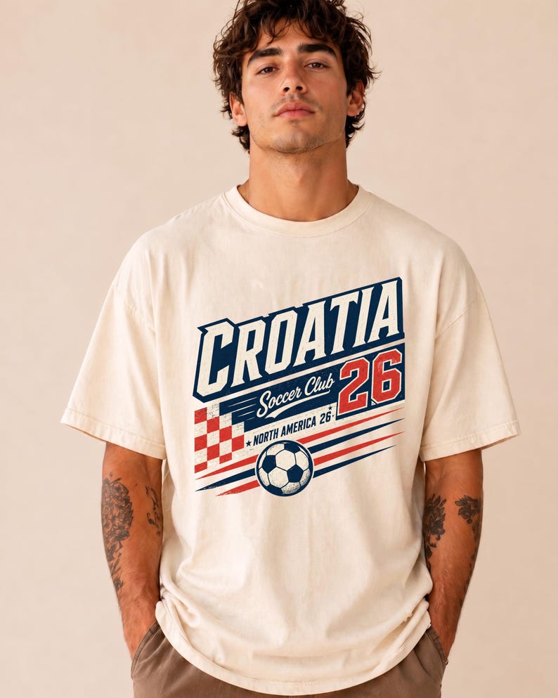 以下が含まれることがあります： CROATIA、Soccer Club、NORTH AMERICA 26という文字がプリントされたクリーム色の半袖Tシャツ。デザインには、赤と白のチェック柄の四角形、赤色の数字26、そして白黒のサッカーボールが含まれています。