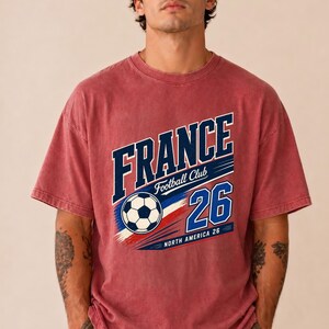 May include: A faded red short-sleeve t-shirt featuring a graphic print that reads FRANCE Football Club 26 NORTH AMERICA 26. The design includes a black and white football, blue and red brushstroke accents, and white lettering.