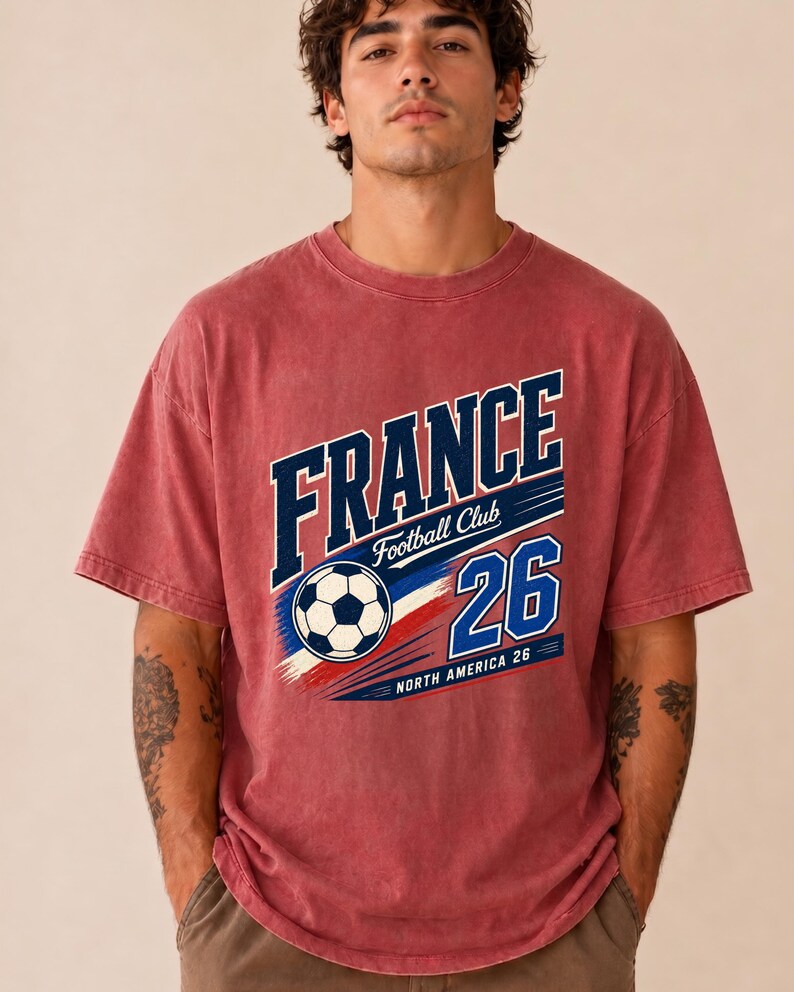 May include: A faded red short-sleeve t-shirt featuring a graphic print that reads FRANCE Football Club 26 NORTH AMERICA 26. The design includes a black and white football, blue and red brushstroke accents, and white lettering.