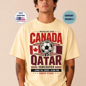 May include: A light yellow t-shirt with a soccer-themed graphic. The design features the text "CANADA VS QATAR" with a soccer ball, flags, and the date "JUNE 18, 2026 | 6:00 PM". The shirt also has the text "VANCOUVER" and "GROUP STAGE".