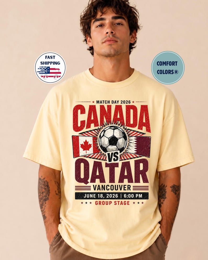 May include: A light yellow t-shirt with a soccer-themed graphic. The design features the text "CANADA VS QATAR" with a soccer ball, flags, and the date "JUNE 18, 2026 | 6:00 PM". The shirt also has the text "VANCOUVER" and "GROUP STAGE".