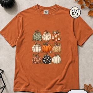 Pode incluir: Uma t-shirt de manga curta cor de laranja queimado com uma impressão gráfica de nove abóboras distintas dispostas numa grelha. As abóboras apresentam vários padrões, incluindo xadrez, bolinhas, motivos florais e cores sólidas, com laços.