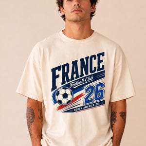 May include: An off-white short-sleeve t-shirt featuring a graphic print with the text FRANCE Football Club and the number 26. The design includes a football and red, white, and blue motion lines, with the text NORTH AMERICA 26 at the bottom.