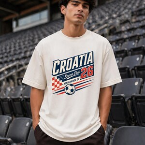 以下が含まれることがあります： 「CROATIA Soccer Club 26」および「NORTH AMERICA 26」の文字がプリントされたオフホワイトの半袖Tシャツ。デザインには赤と白の市松模様、サッカーボール、赤と青の水平線が含まれています。