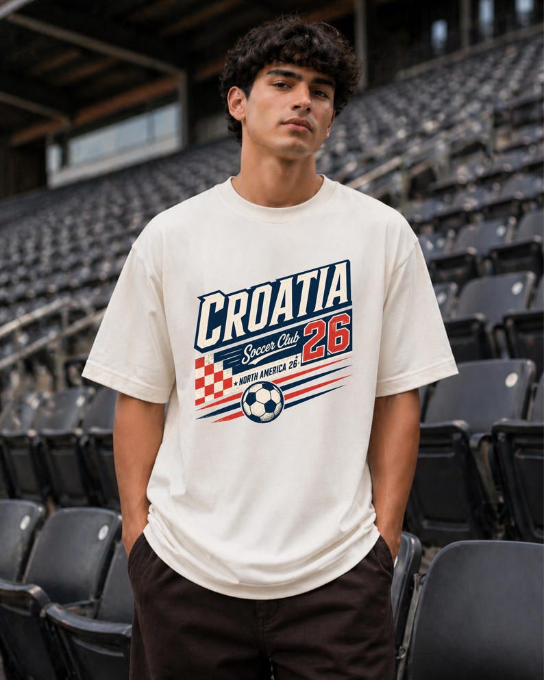 以下が含まれることがあります： 「CROATIA Soccer Club 26」および「NORTH AMERICA 26」の文字がプリントされたオフホワイトの半袖Tシャツ。デザインには赤と白の市松模様、サッカーボール、赤と青の水平線が含まれています。