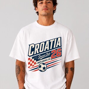 以下が含まれることがあります： CROATIA、Soccer Club、26、NORTH AMERICA 26というテキストがプリントされた白い半袖Tシャツ。デザインには、赤と白の市松模様の正方形、サッカーボール、赤と青の水平線が含まれています。