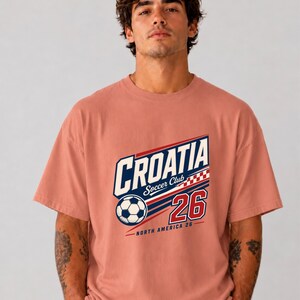 K&ouml;nnte beinhalten: Ein altrosa T-Shirt mit einem Grafikdruck auf der Brust, der den Text CROATIA Soccer Club, einen Fu&szlig;ball, ein rot-wei&szlig;es Karomuster, die Zahl 26 sowie den Text NORTH AMERICA 26 in blauer, wei&szlig;er und roter Farbe zeigt.