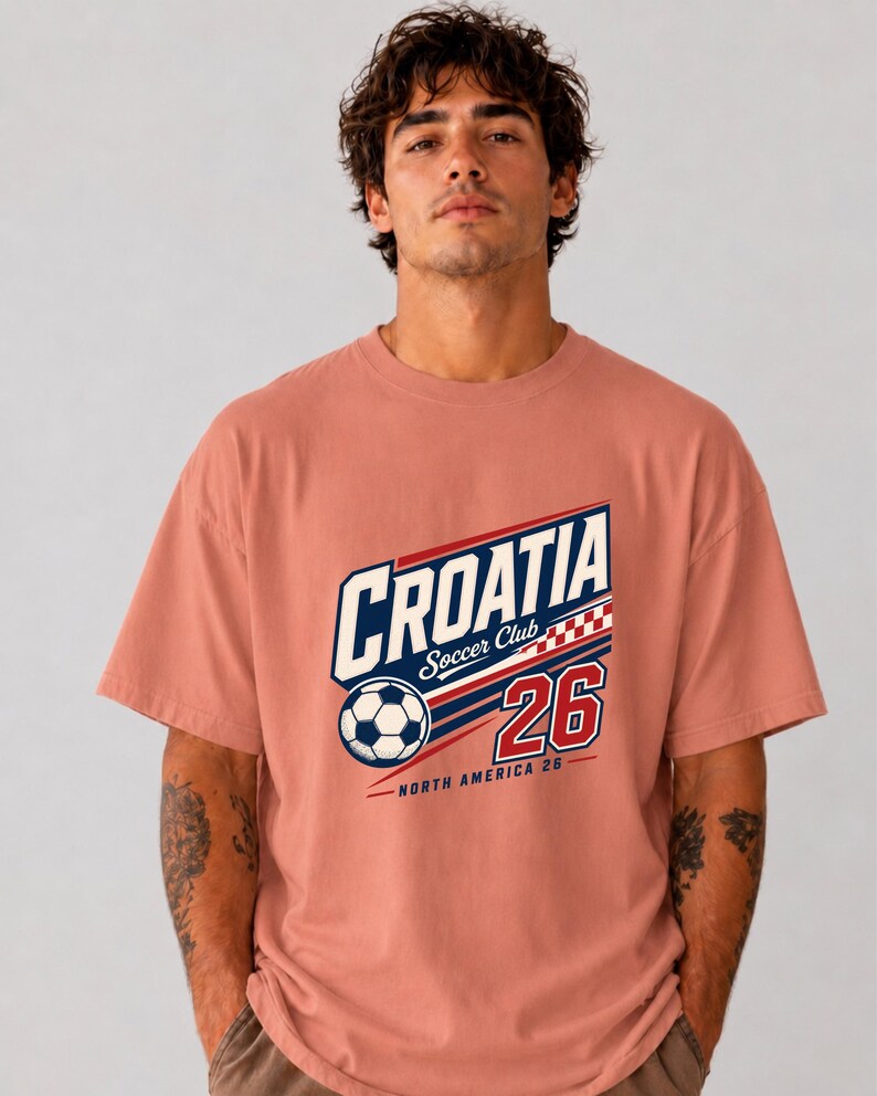 K&ouml;nnte beinhalten: Ein altrosa T-Shirt mit einem Grafikdruck auf der Brust, der den Text CROATIA Soccer Club, einen Fu&szlig;ball, ein rot-wei&szlig;es Karomuster, die Zahl 26 sowie den Text NORTH AMERICA 26 in blauer, wei&szlig;er und roter Farbe zeigt.
