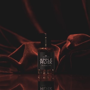 May include: A perfume bottle with a black label that reads "APPLE MUSK Perfume Oil" is centered in front of a dark red, draped fabric. The bottle is clear, and the liquid inside is amber-colored. The background is a deep red.