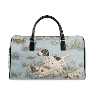 May include: A light blue duffel bag featuring a hunting scene with dogs and a duck. The bag has black handles and accents, a gold zipper, and a detailed illustration of dogs in a field with tall grass. The bag is designed for travel.
