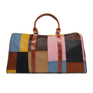 Può includere: Una borsa da viaggio patchwork multicolore con manico e cinghie in pelle marrone. La borsa presenta pannelli rettangolari in vari colori, tra cui blu, giallo, rosa, nero e marrone. La borsa è progettata per i viaggi e ha una chiusura a cerniera.