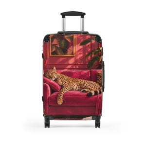 May include: A rolling suitcase featuring a printed graphic of a cheetah sleeping on a vibrant magenta velvet armchair. The luggage has a black frame, a retractable handle, and four wheels. The background print includes a framed botanical art piece.