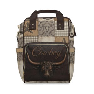 Könnte beinhalten: Ein Rucksack mit Patchwork-Design in Beige-, Braun- und Grautönen. Die Vordertasche zeigt ein dunkelbraunes, strukturiertes Feld mit dem Wort Cowboy in Schreibschrift und einer Stierkopf-Illustration. Er hat zwei dunkelbraune Tragegriffe.