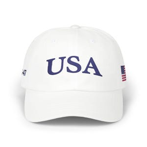 May include: White baseball cap with the word "USA" in navy blue script across the front. A small American flag is on the side, and the number "47" is on the other side. The cap has a curved brim.