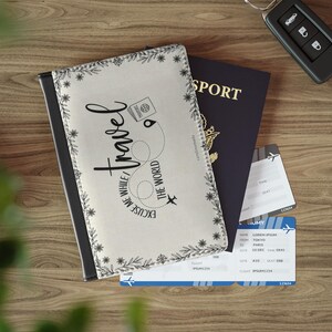 以下が含まれることがあります： 「Travel, Excuse Me While I See The World」の黒い文字が書かれたクリーム色のパスポートカバー。パスポートと航空券も表示されています。カバーは黒い縁取りと花のアクセントが付いています。