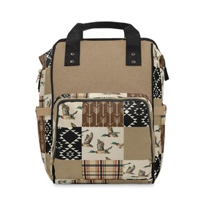 May include: A backpack featuring a patchwork design with tan, brown, black, and white colours. The pattern includes flying mallard ducks, white arrows on brown squares, black and white geometric shapes, and a tan, black, and red plaid section.