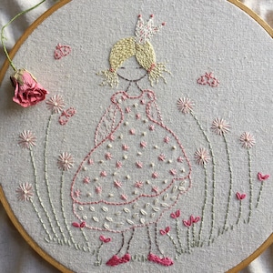 Pode incluir: Arte de bordado em aro que mostra uma menina usando um vestido rosa e uma coroa, em pé em um campo de flores. As flores são rosa, brancas e verdes.