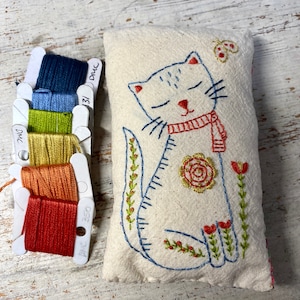 Könnte beinhalten: Ein weißes Kissen mit einer handgestickten Stickerei eines blauen Katers mit einem roten Schal. Die Katze ist von grünen und roten Blumen und Blättern umgeben. Das Kissen ist mit DMC-Stickgarn verziert.