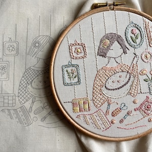 May include: An embroidered artwork in a wooden hoop, depicting a woman surrounded by framed floral designs and craft supplies. The colour palette includes soft pastels like pink, peach, and green. The design also features a sketch outline, giving it a handmade feel.
