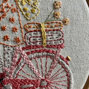 May include: Close-up of a pink and white embroidered bicycle with a basket filled with yellow and orange flowers. The bicycle is on a white fabric background.