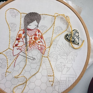 Peut inclure: Une broderie représentant une personne assise dans un fauteuil, portant une robe à motifs colorés. L'œuvre est brodée sur un tissu blanc avec un fil jaune. L'arrière-plan présente des motifs floraux et une petite plante en pot.