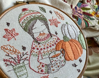 May include: Embroidered hoop art featuring a person wearing a brown and green hat and sweater holding a mug of hot beverage. The person is surrounded by fall leaves and pumpkins.