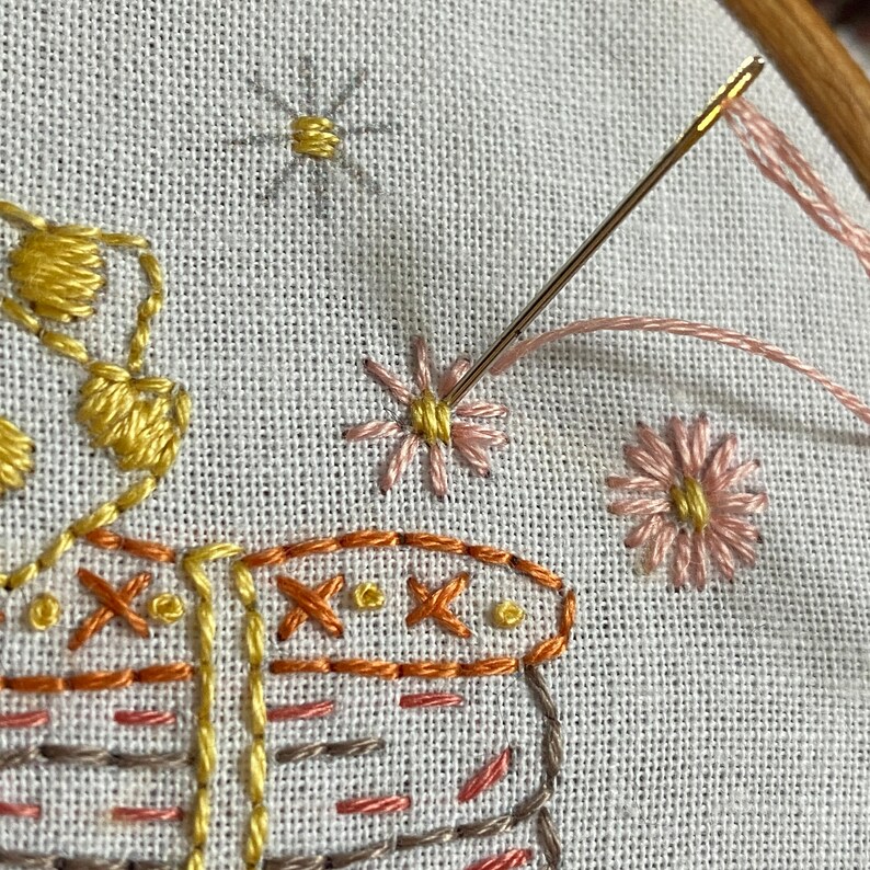 May include: Close-up of embroidery on white fabric. A pink flower with a yellow center is being stitched with a gold needle and pink thread. Other embroidered details include a yellow flower, a star, and a brown and orange patterned design.