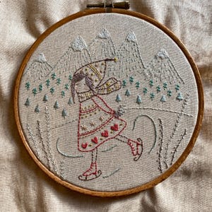 Pode incluir: Arte de bordado em aro que representa uma menina patinando no gelo em uma paisagem nevada. A menina está usando um vestido vermelho e branco com um chapéu e uma echarpe marrons. O fundo é uma paisagem nevada com montanhas e árvores.