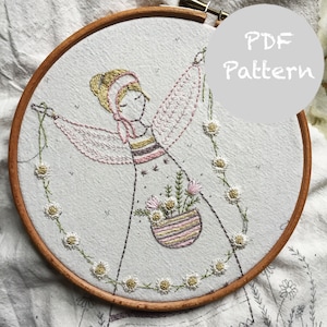 Pode incluir: Arte de bordado em um aro que representa uma menina com uma cesta de flores e uma corrente de margaridas. O aro é de madeira e o bordado é feito em tons de rosa, branco e verde. O texto "PDF Pattern" é visível no canto superior direito da imagem.