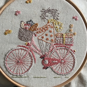 May include: Embroidered artwork of a person riding a pink bicycle with a basket full of items. The person is wearing a patterned dress and a floral headband. The background is a light beige fabric.
