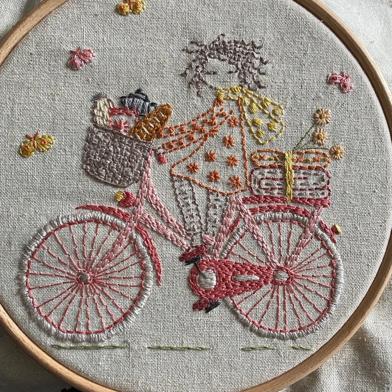 May include: Embroidered artwork of a person riding a pink bicycle with a basket full of items. The person is wearing a patterned dress and a floral headband. The background is a light beige fabric.