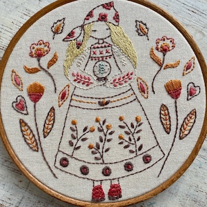 May include: Embroidered folk art in a wooden hoop. The design shows a stylized figure with blonde hair, wearing a dress and head covering, surrounded by floral and leaf motifs in red, orange, and brown. The embroidery is on a cream-coloured fabric.