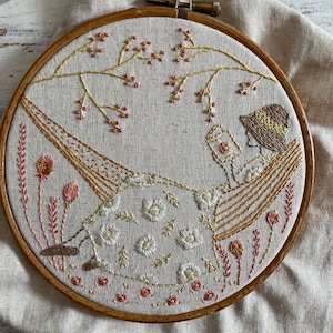 Pode incluir: Arte de bordado em aro que mostra uma pessoa relaxando em uma rede com um livro, cercada por detalhes florais delicados em tons de rosa, amarelo e branco.