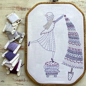 Pode incluir: Um aro de bordado branco com um desenho bordado em roxo e branco de uma menina segurando uma árvore de Natal. A menina está usando um chapéu branco e um vestido roxo. A árvore está decorada com listras roxas e brancas. O bordado está sobre um fundo de tecido branco.