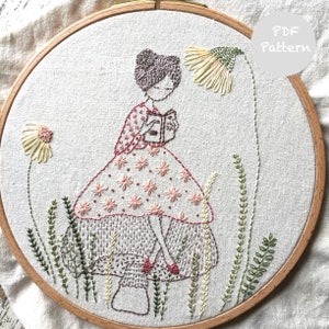 Pode incluir: Arte de bordado em aro com uma menina em um vestido floral rosa e branco lendo um livro enquanto está sentada em um cogumelo. O fundo inclui grama verde e flores amarelas.