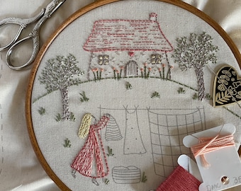 Peut inclure: Un cerceau de broderie fait main représentant un chalet avec un toit rose, une fille en robe rouge et une corde à linge avec du linge suspendu. Le cerceau est sur un fond de lin blanc.