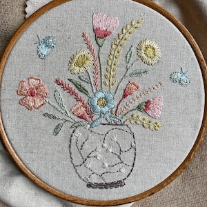 May include: A hand-stitched embroidery hoop featuring a bouquet of flowers in a cream-colored vase. The flowers are pink, yellow, blue, and green. The embroidery is on a beige linen fabric.