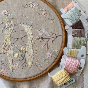 Peut inclure: Gros plan d'un cerceau de broderie cousu à la main avec un motif représentant une fille aux longs cheveux blonds et un oiseau sur une branche. La broderie est réalisée dans des tons de rose, vert et jaune. Le cerceau est entouré de plusieurs bobines de fil à broder de différentes couleurs.