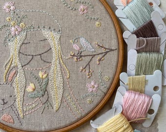 Peut inclure: Gros plan d'un cerceau de broderie cousu à la main avec un motif représentant une fille aux longs cheveux blonds et un oiseau sur une branche. La broderie est réalisée dans des tons de rose, vert et jaune. Le cerceau est entouré de plusieurs bobines de fil à broder de différentes couleurs.