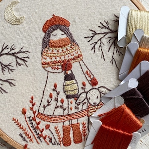Può includere: Opera d'arte ricamata in un telaio di legno. Il disegno presenta una figura con berretto e sciarpa arancioni, che tiene una lanterna ed è accompagnata da un agnello. La tavolozza dei colori comprende arancione, marrone e beige, con una luna crescente e rami sullo sfondo.