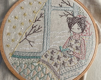 May include: Embroidered hoop art featuring a person sitting by a window, reading a book. The scene is decorated with a floral pattern and a whimsical design of a tree with branches reaching up to the top of the hoop.