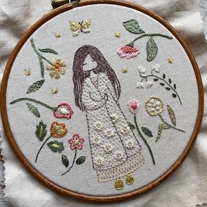 Pode incluir: Um bastidor de madeira circular emoldura um tecido cinza-claro com um desenho bordado à mão de uma pessoa com longos cabelos castanhos vestindo um vestido branco adornado com pequenas flores amarelas e brancas, rodeado por várias flores bordadas coloridas e uma pequena borboleta amarela.